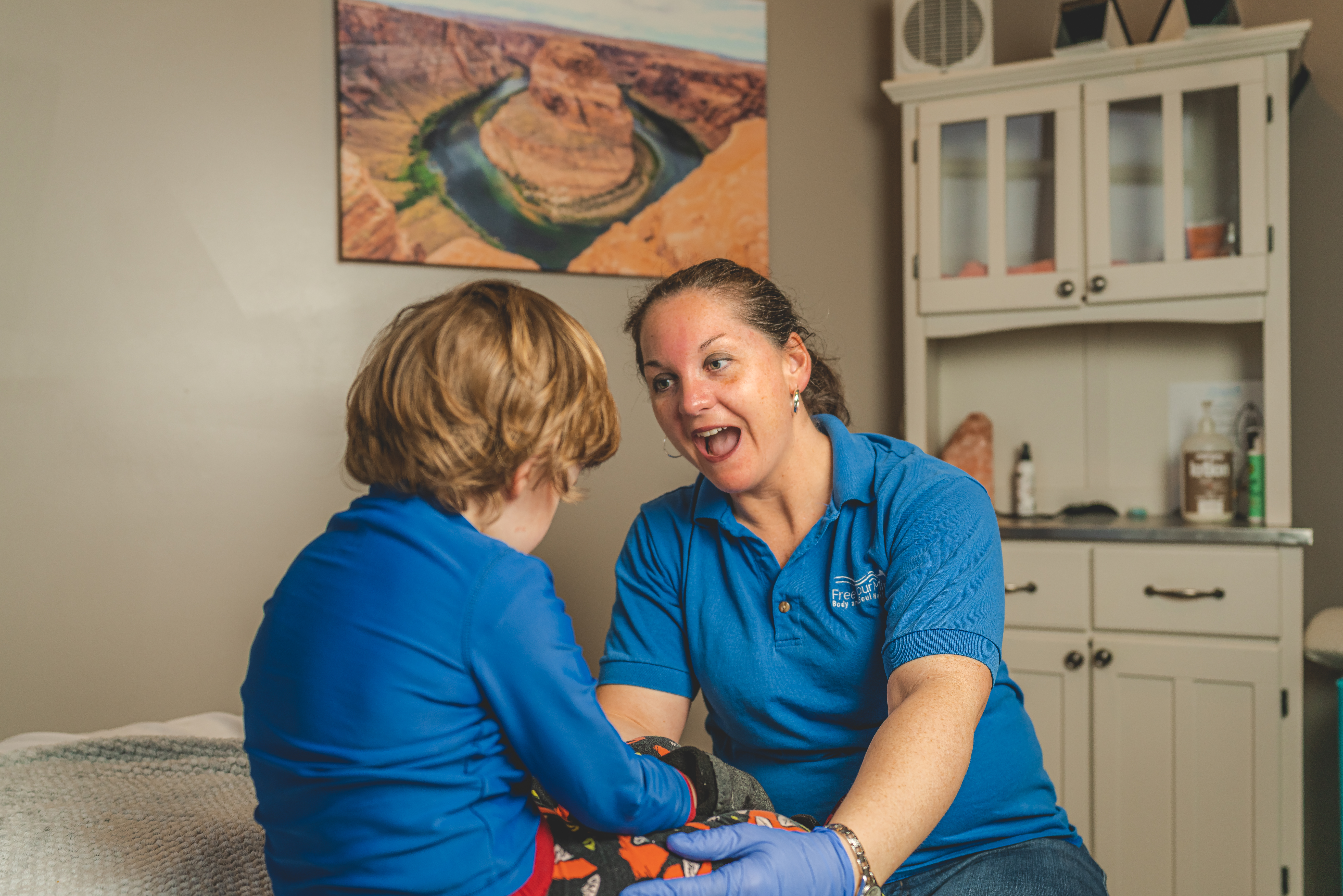 Healthcare worker in a blue polo smiles warmly while assisting a child in blue clothing; canyon artwork hangs in the background.