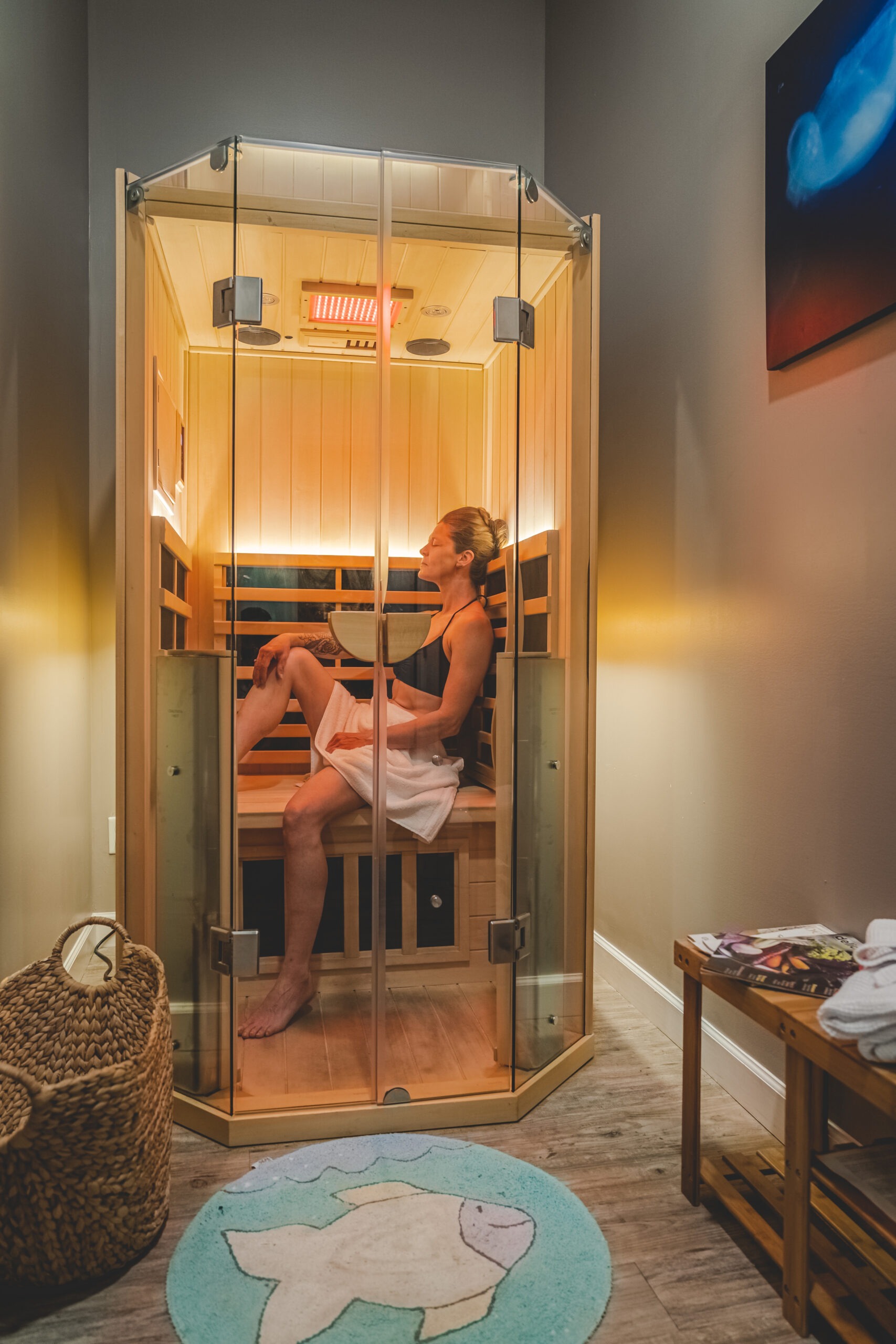 A woman relaxes in a glass-enclosed sauna, draped in a towel, highlighting wellness and self-care. A fish-patterned rug and towels are nearby.