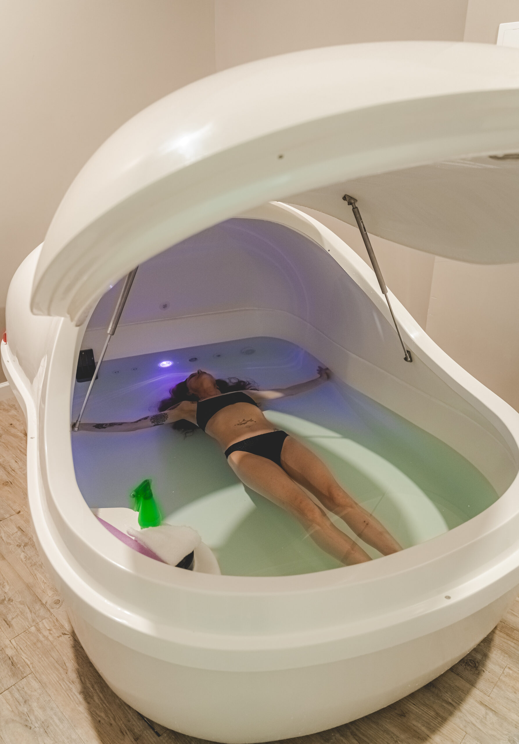 A woman floats in a sensory deprivation tank, surrounded by light blue water. The serene environment emphasizes relaxation and wellness.