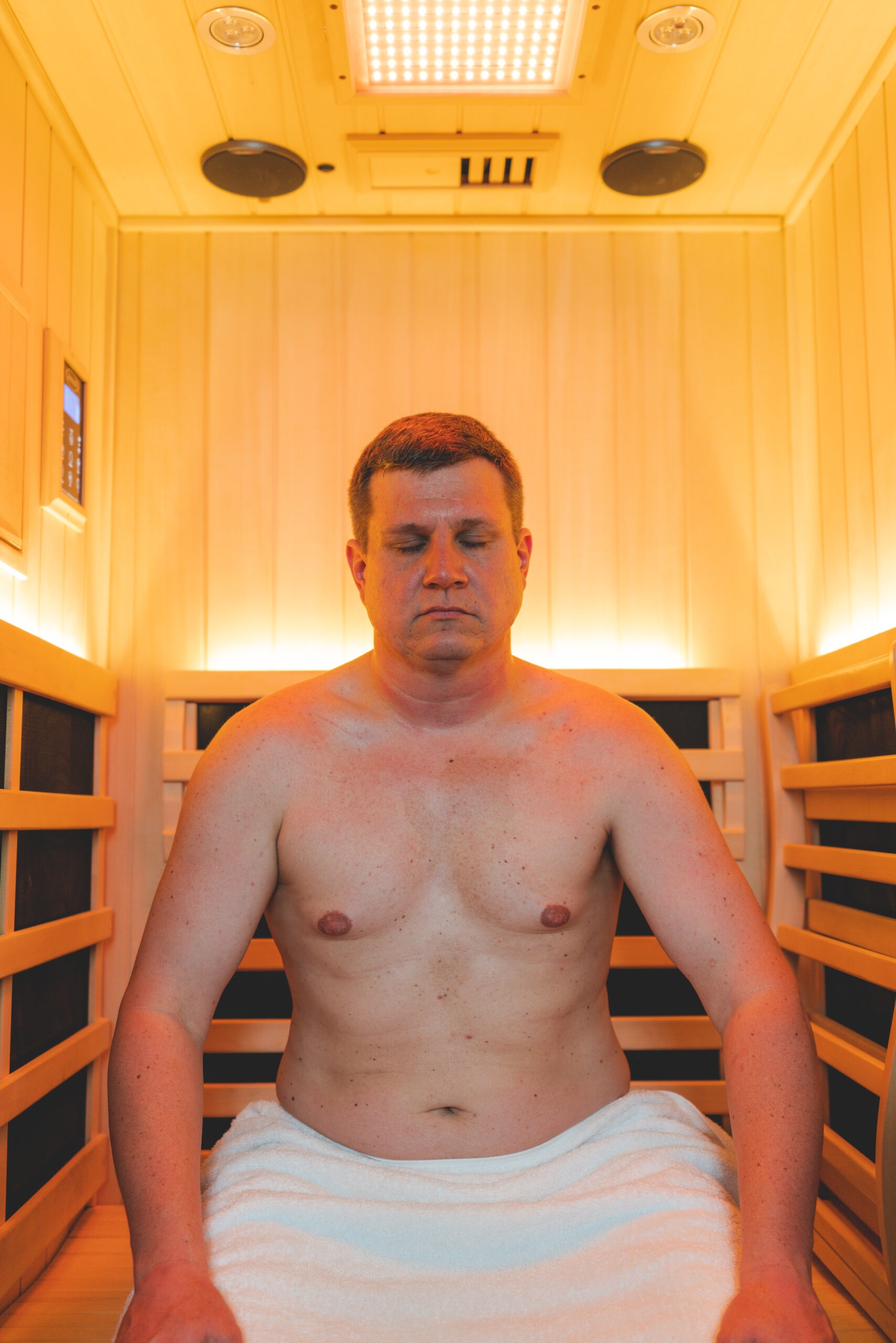 A man sitting in a wooden infrared sauna, eyes closed, wearing a towel, appearing relaxed amidst warm lighting. The image highlights wellness and relaxation.