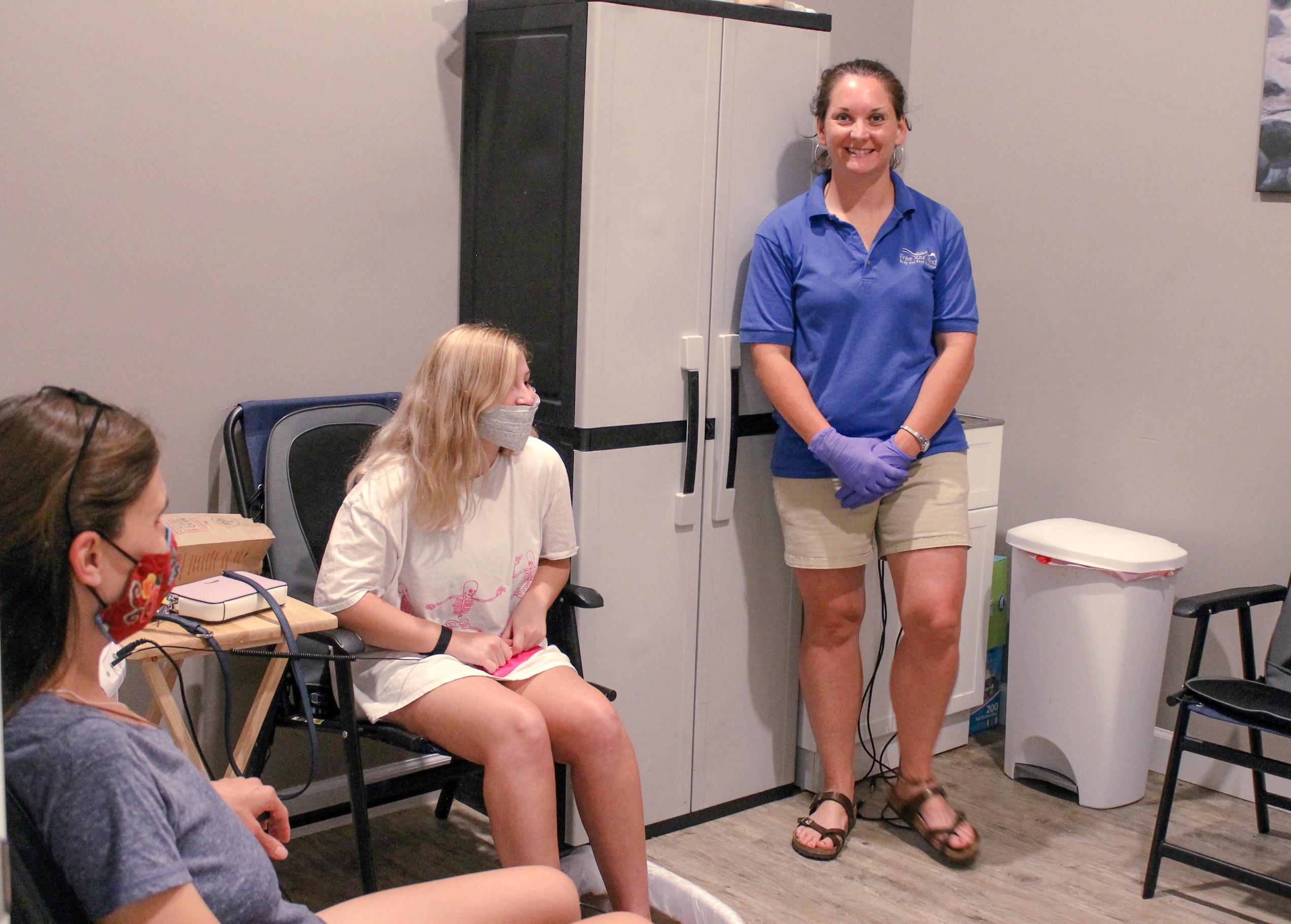 A healthcare worker in a blue shirt stands beside two patients in a waiting area, both wearing masks. The setting suggests a medical consultation.