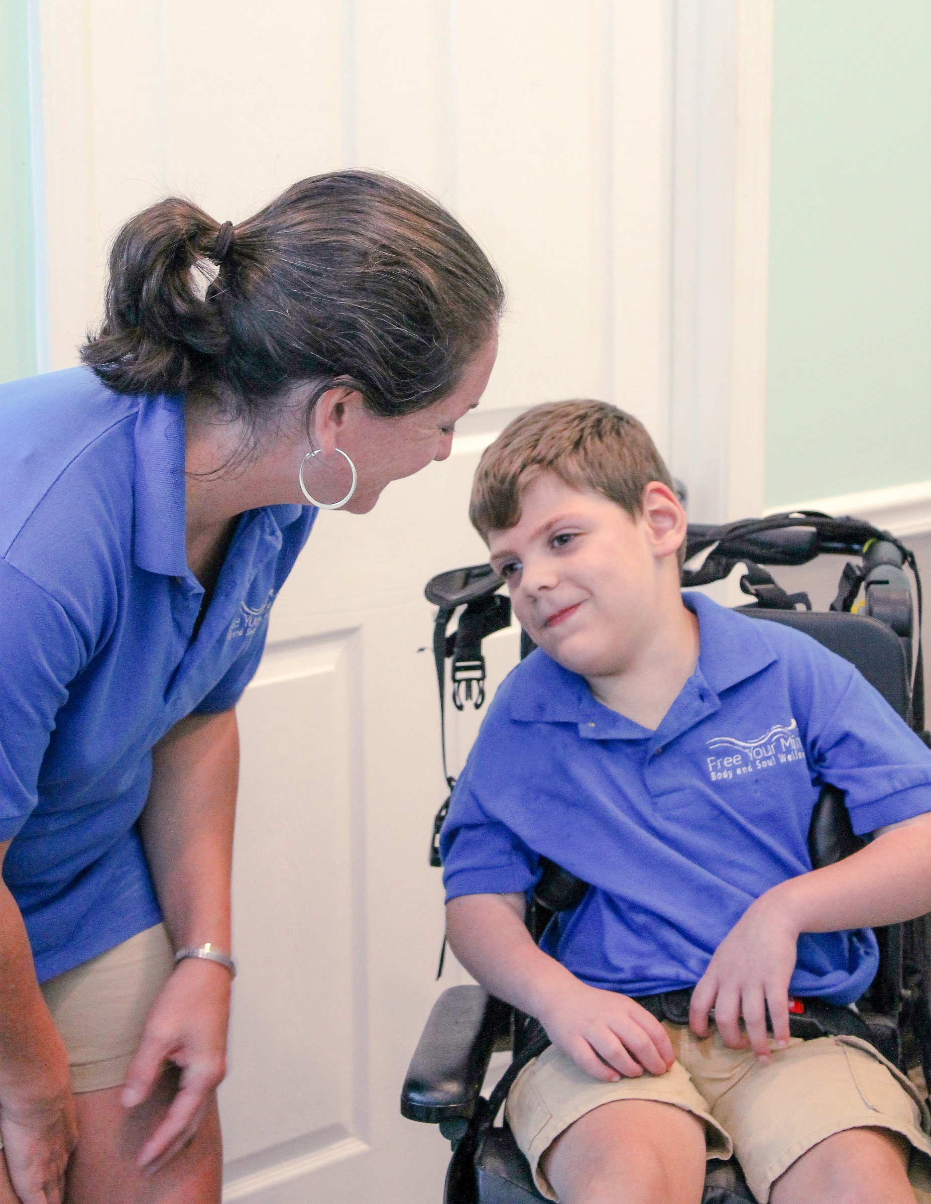 A young boy in a wheelchair smiles at a woman in a blue shirt during a warm interaction, emphasizing support and connection.