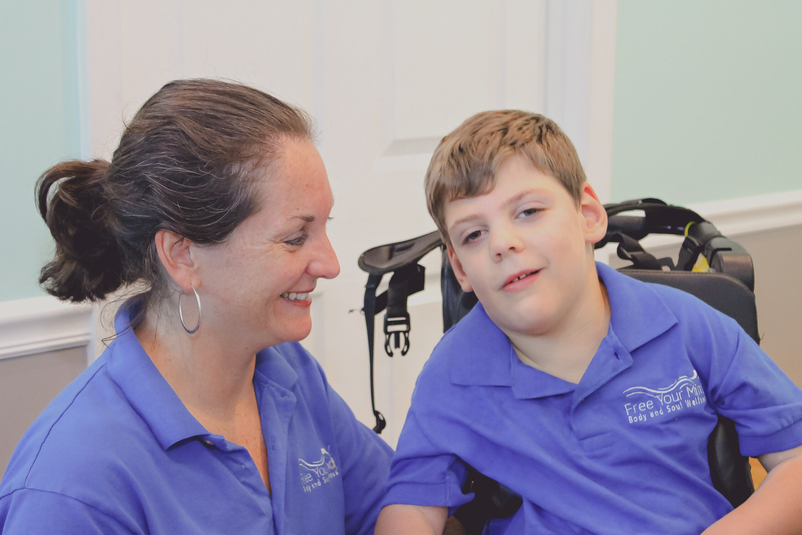 A smiling woman and a boy in a wheelchair share a joyful interaction, both wearing blue shirts featuring "Free Your Mind." This image highlights supportive connections in a caring environment.