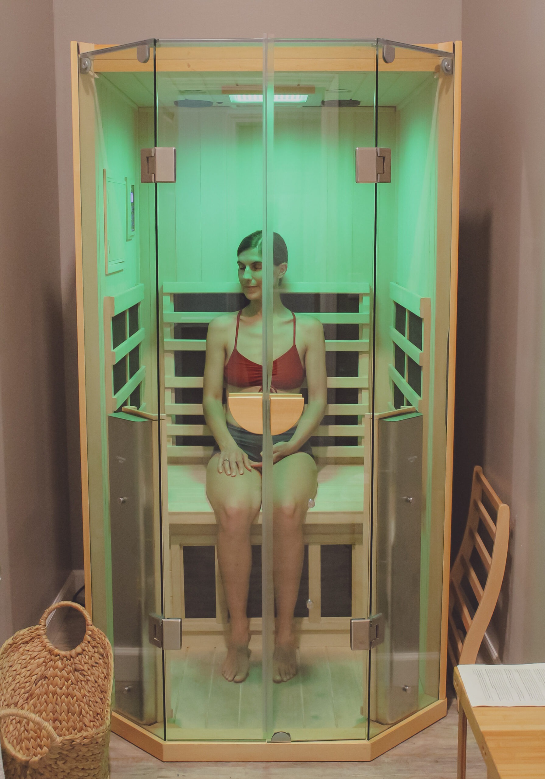 A woman sits relaxed in a glass sauna emitting green light, promoting wellness and relaxation. A basket and chair are visible nearby.