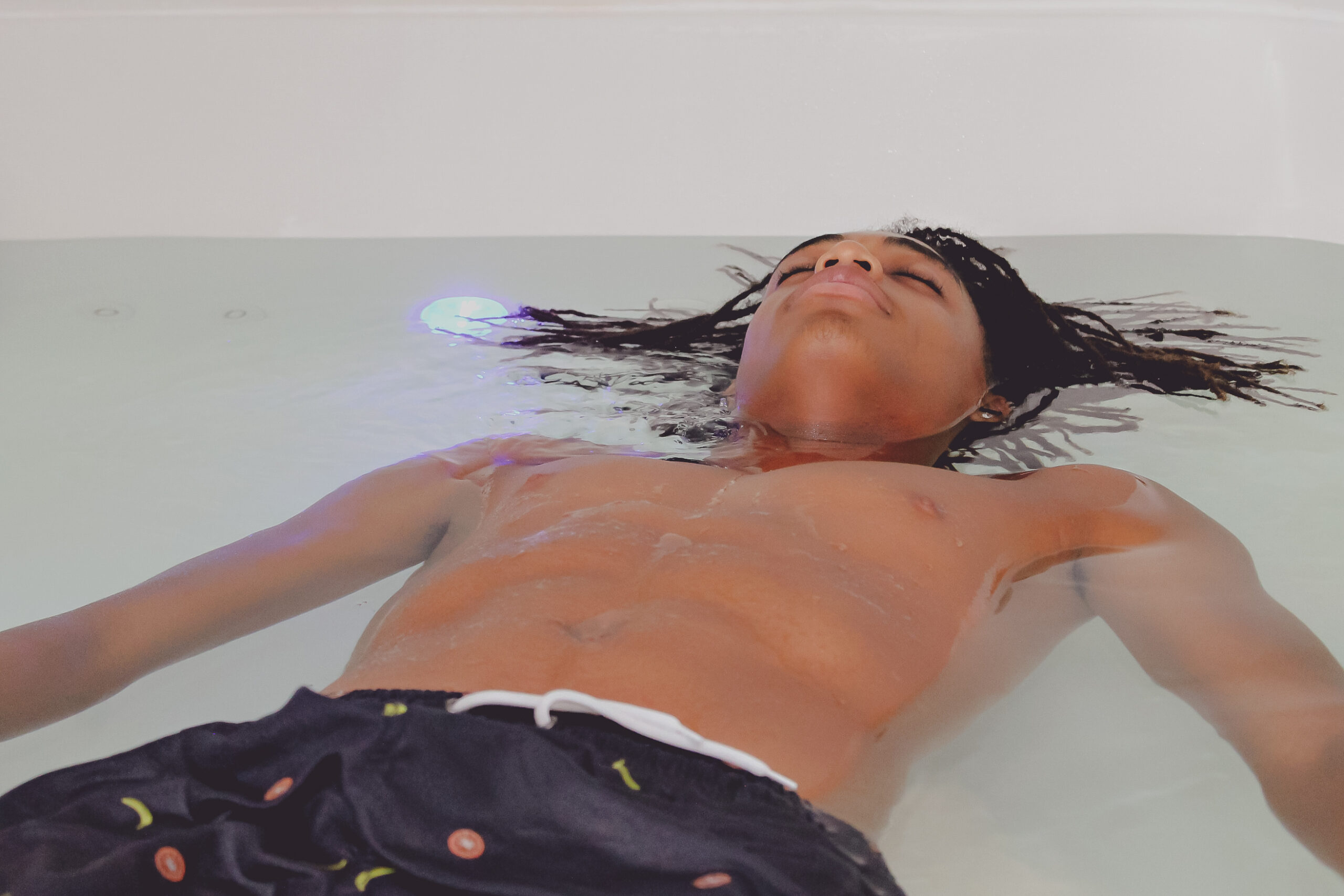 Teenager reclining in a bath with serene expression, water gently rippling around him, showcasing relaxation and tranquility.